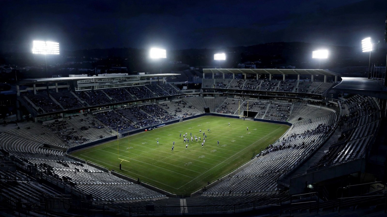 An American football stadium lit by floodlights at night, with players on the field and spectators in the stands