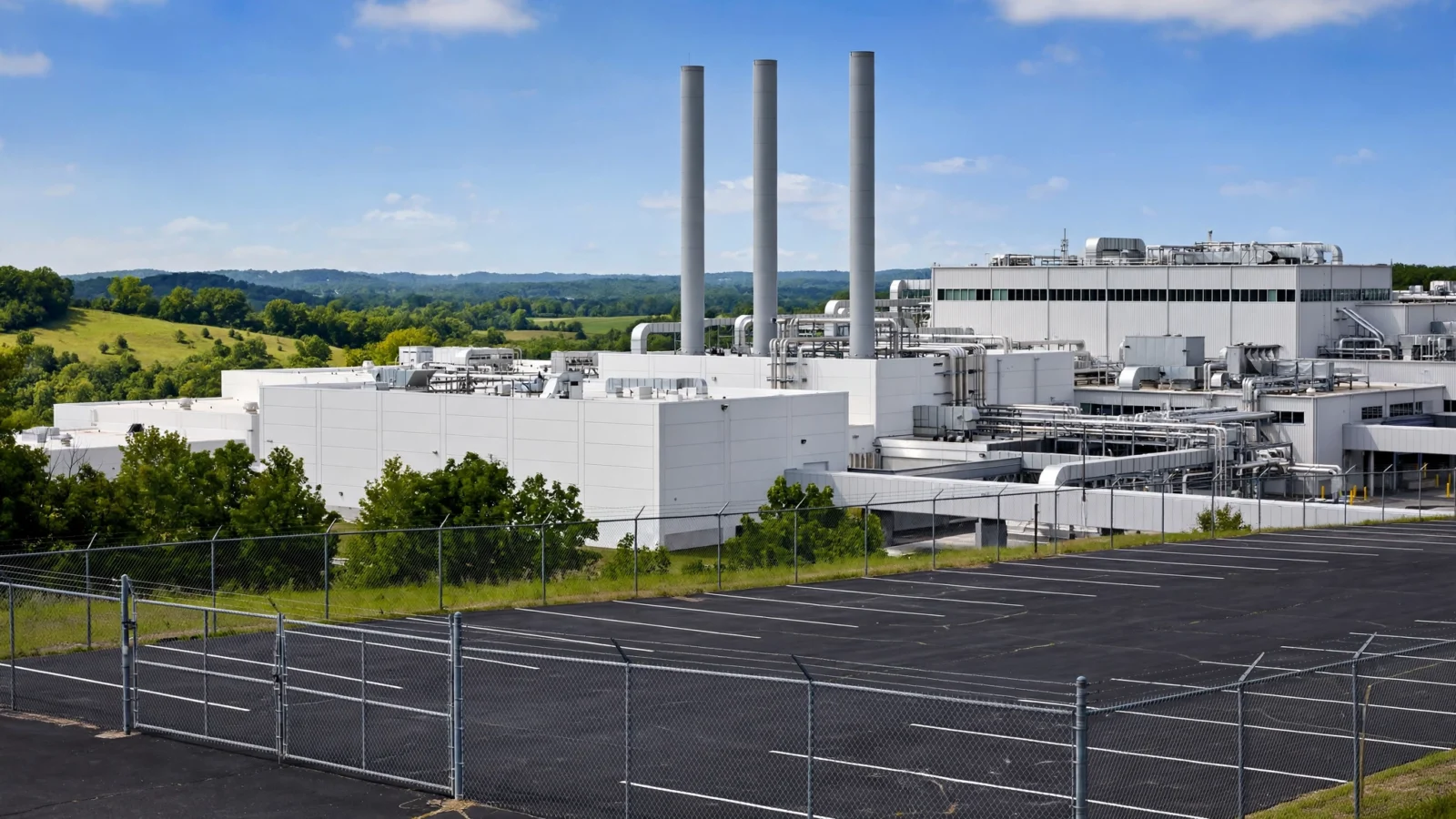 Exterior of a large white pharmaceutical manufacturing plant with tall smokestacks behind an empty parking lot, set against green hills under a blue sky.