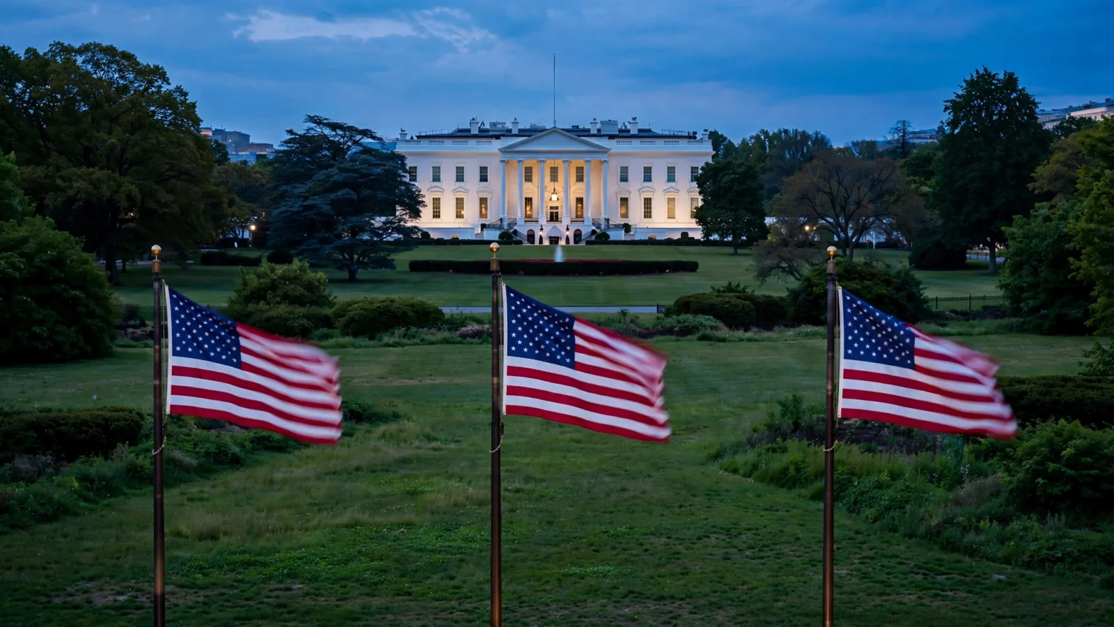 The White House exterior lit at dusk, seen across the lawn with three American flags flying in the foreground.
