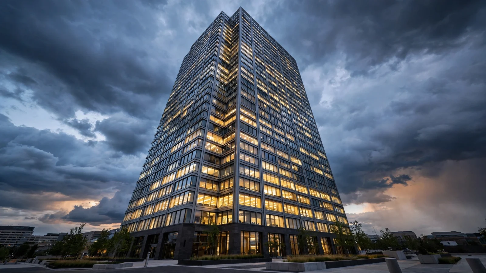 A modern glass office tower photographed from below at dusk, its windows lit against a cloudy evening sky.