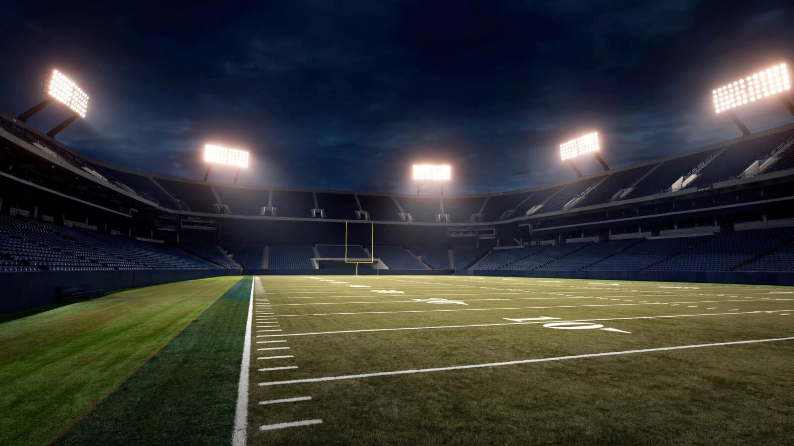 An empty American football stadium lit by floodlights at night, with the field, yard lines and a goalpost in the distance.