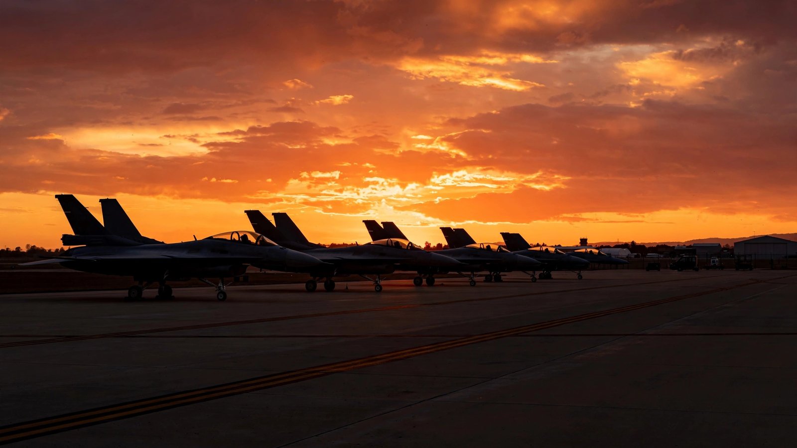 Fighter jets parked on an airbase runway silhouetted against an orange sunset