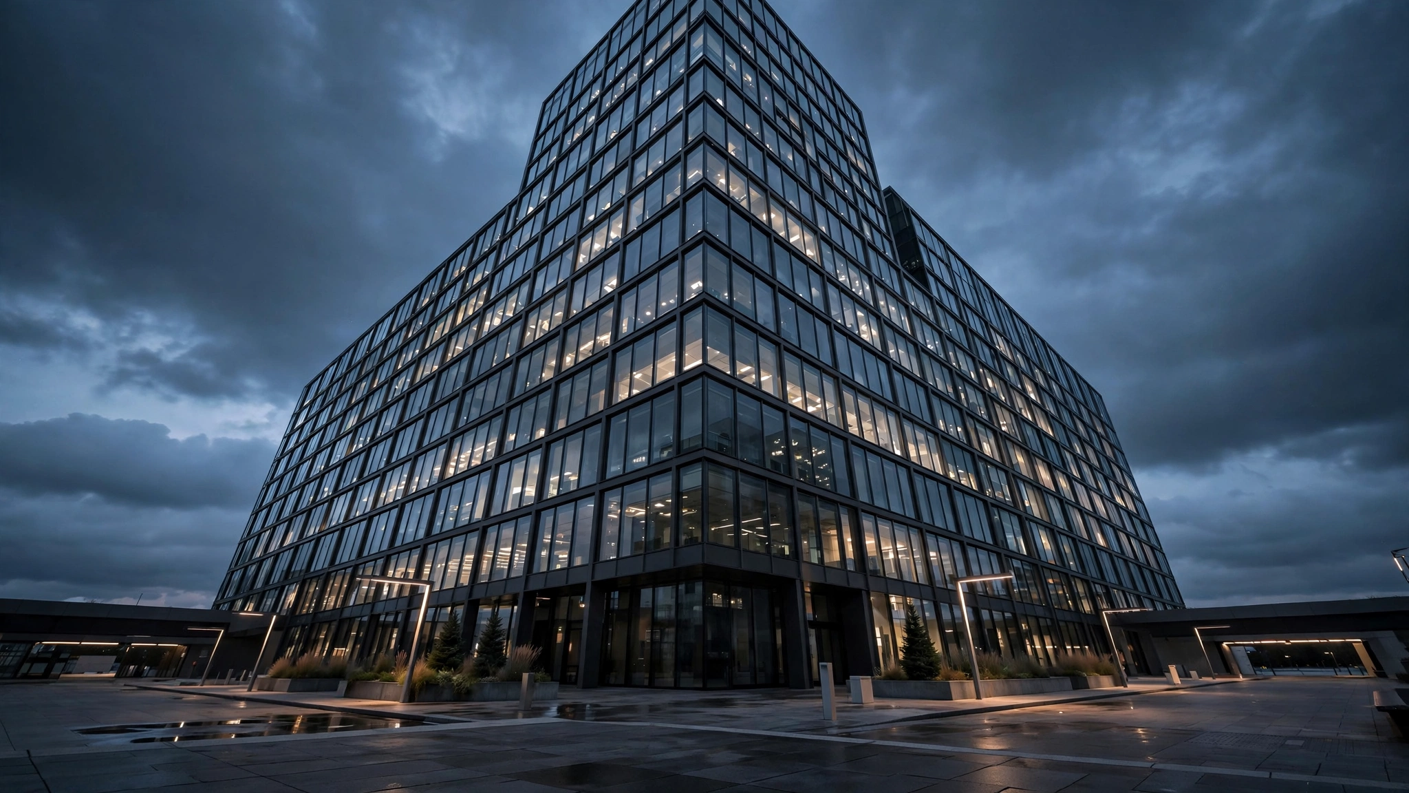 A modern glass corporate office building with lit windows photographed from the plaza at dusk under dark clouds