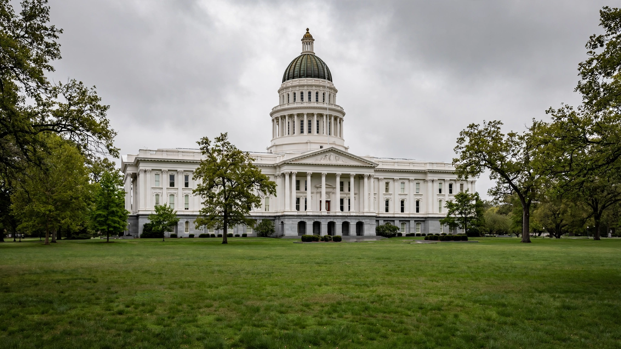 A domed state capitol building with white columns seen from across a green lawn under a cloudy sky