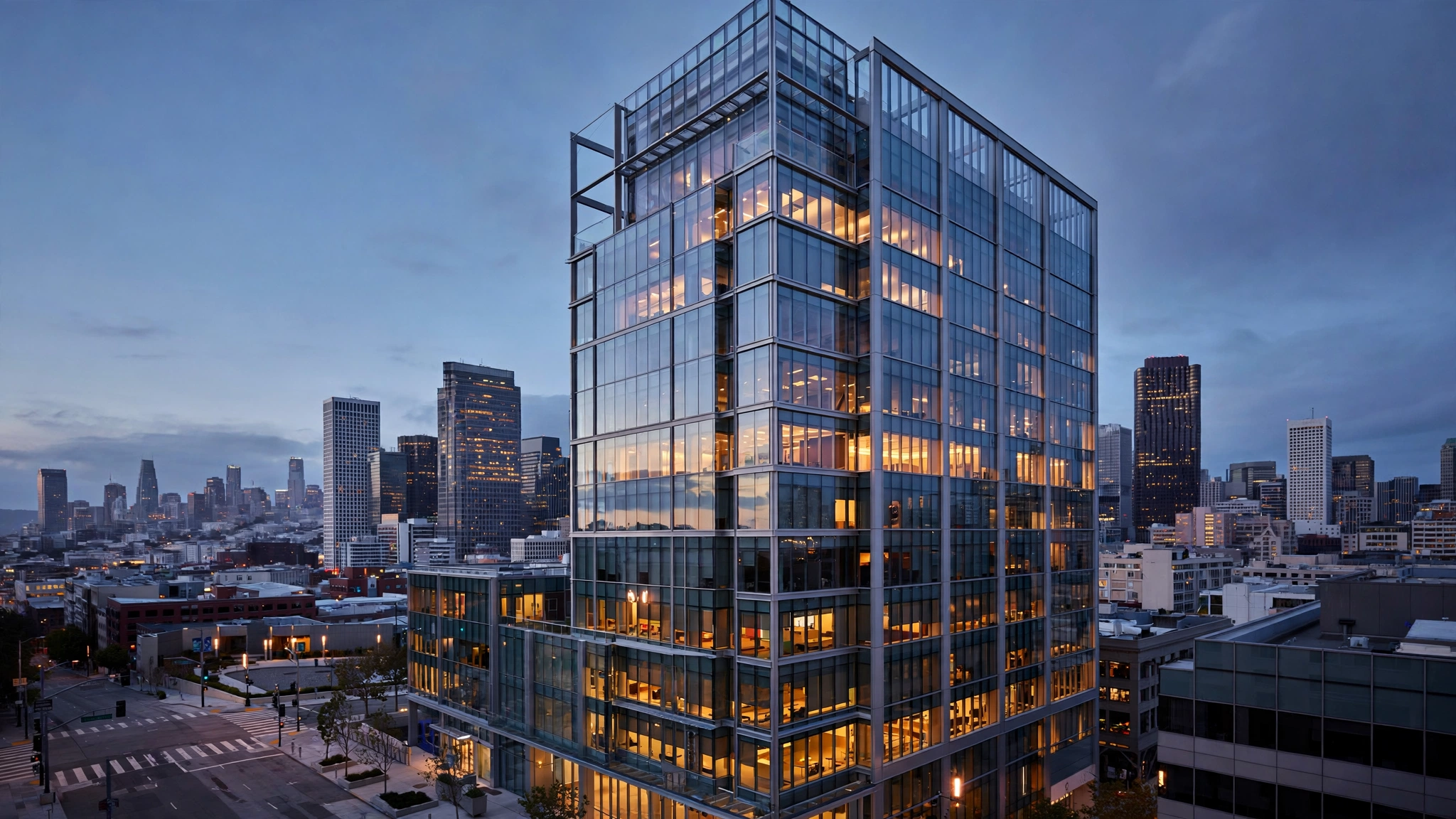 A glass office tower with lit windows at dusk in a downtown skyline