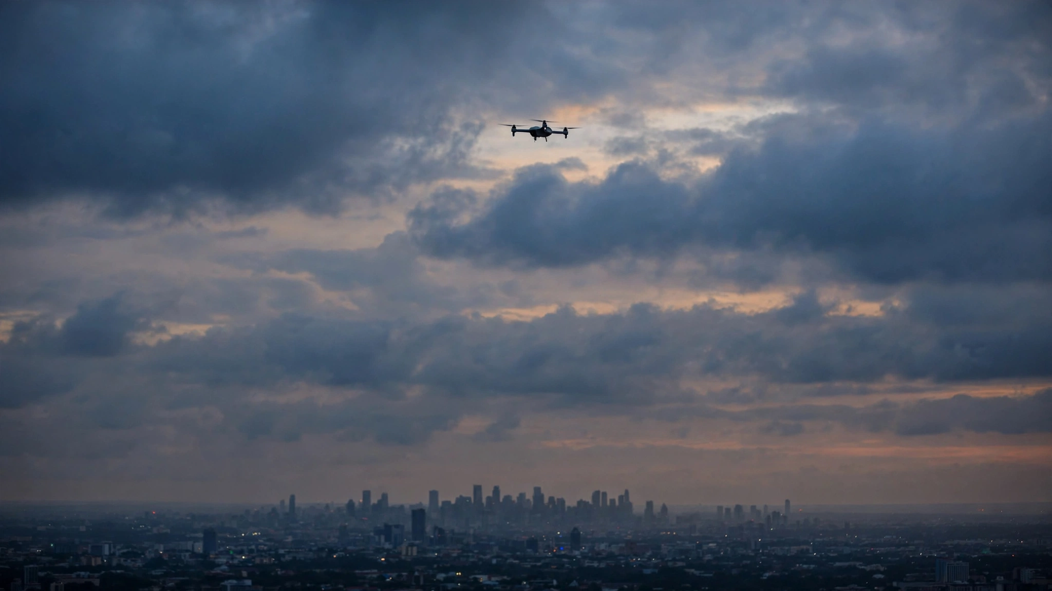 A small drone flying in a cloudy dusk sky above a distant city skyline
