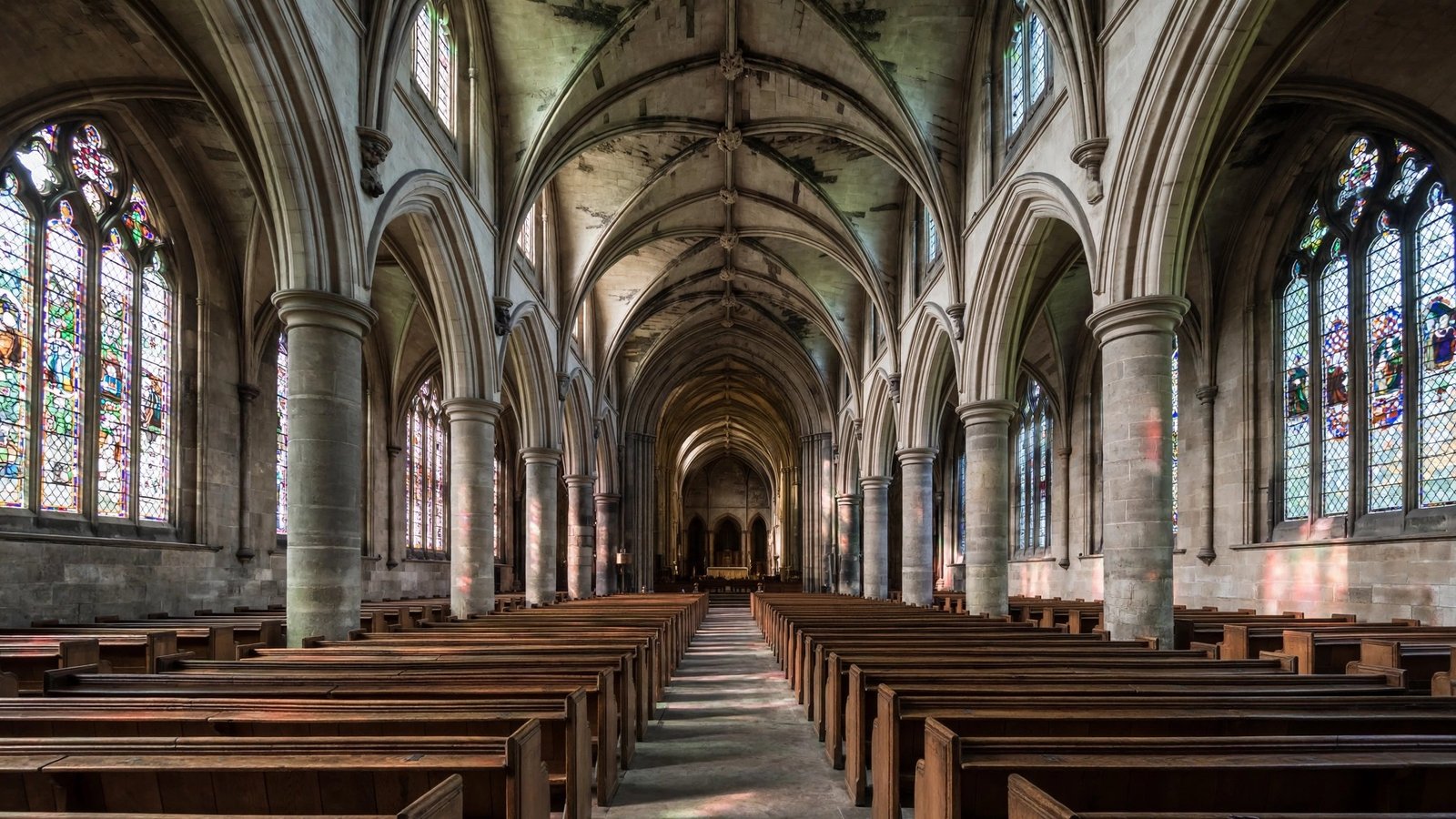 The empty nave of a cathedral with stone columns, an arched ceiling, stained glass windows and rows of wooden pews