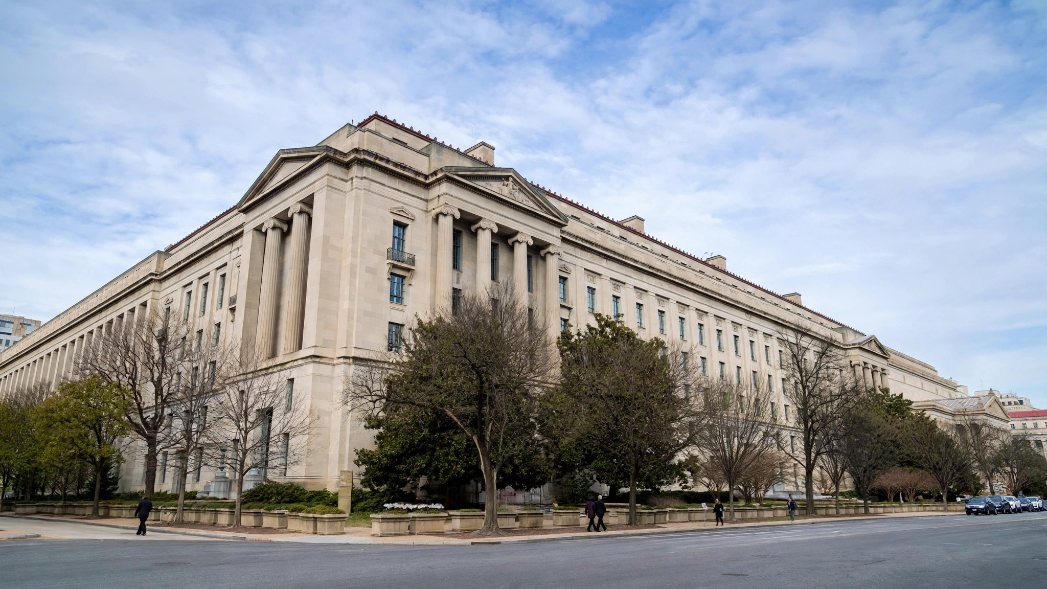 The stone facade of the United States Department of Justice building in Washington, with trees and a street in front