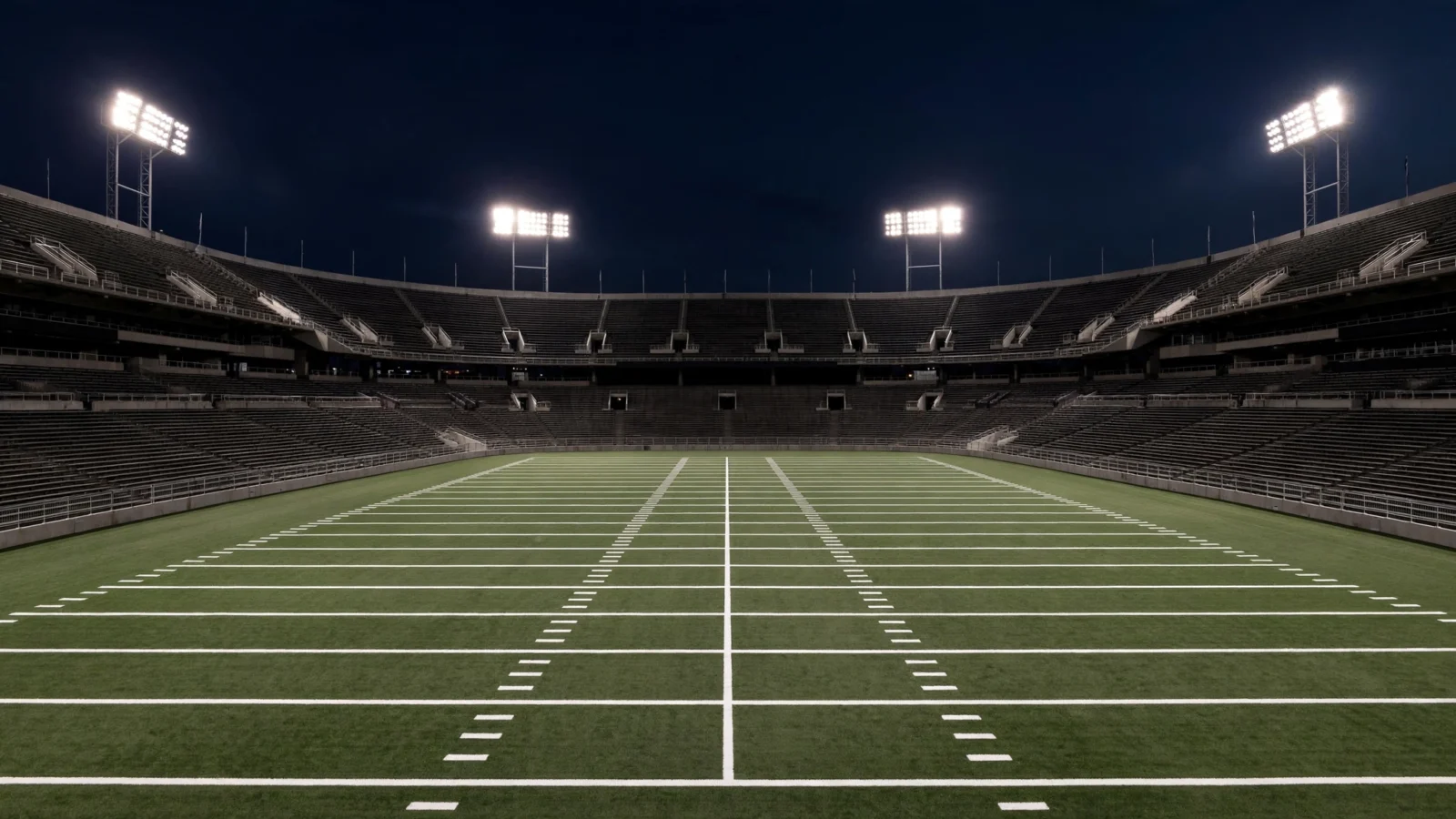 An empty American football stadium under floodlights at night, with yard lines marked on the field