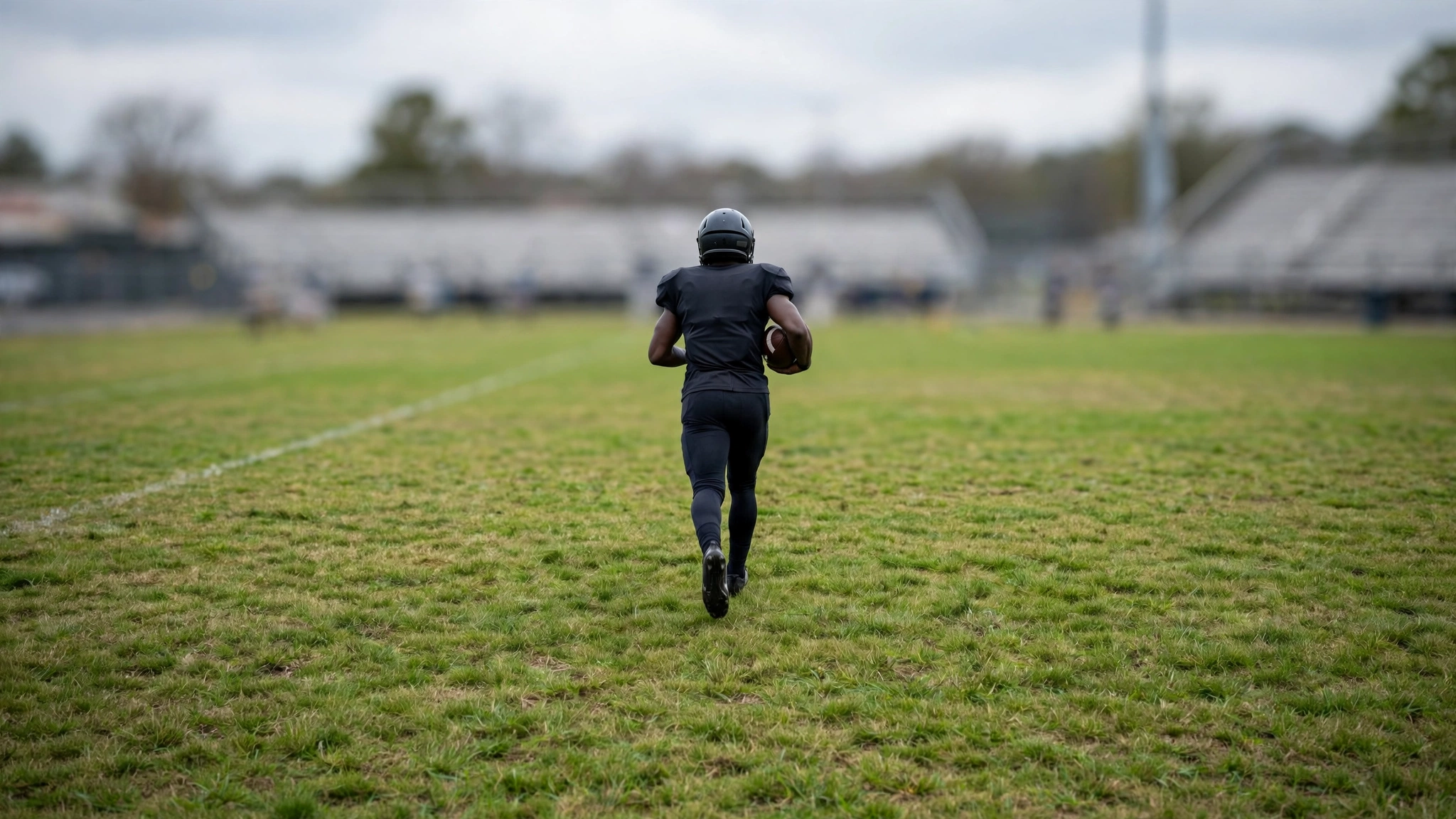 A football player in a plain dark uniform running with a football on a grass field, seen from behind