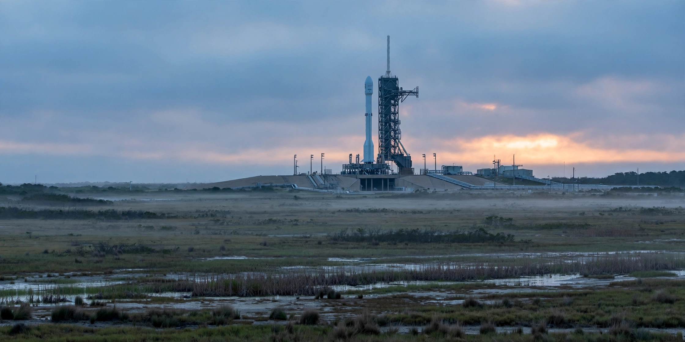 A white rocket standing beside a launch tower on a flat launch pad at dawn under a cloudy sky