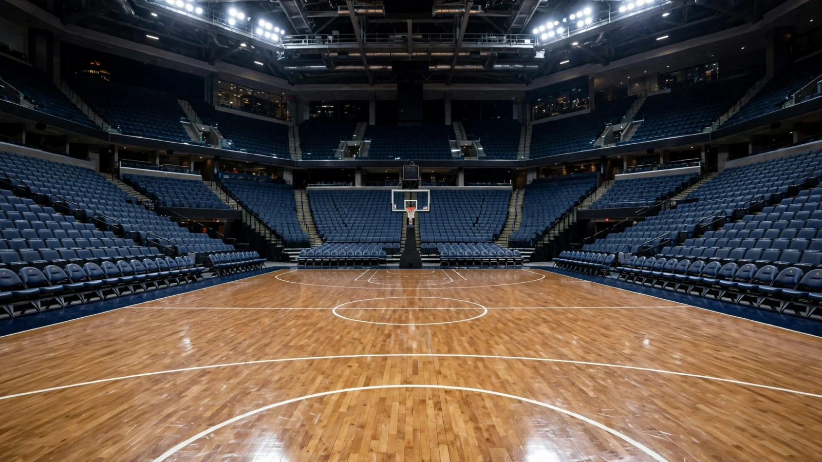 Empty basketball arena with a polished wooden court, a hoop, and rows of blue seats under bright lights