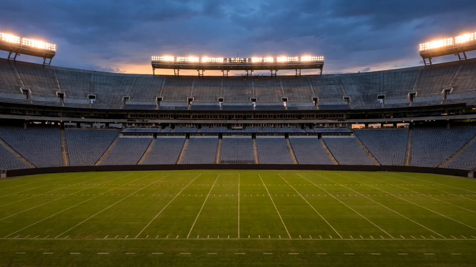 An empty American football stadium under floodlights at dusk, with a marked green field