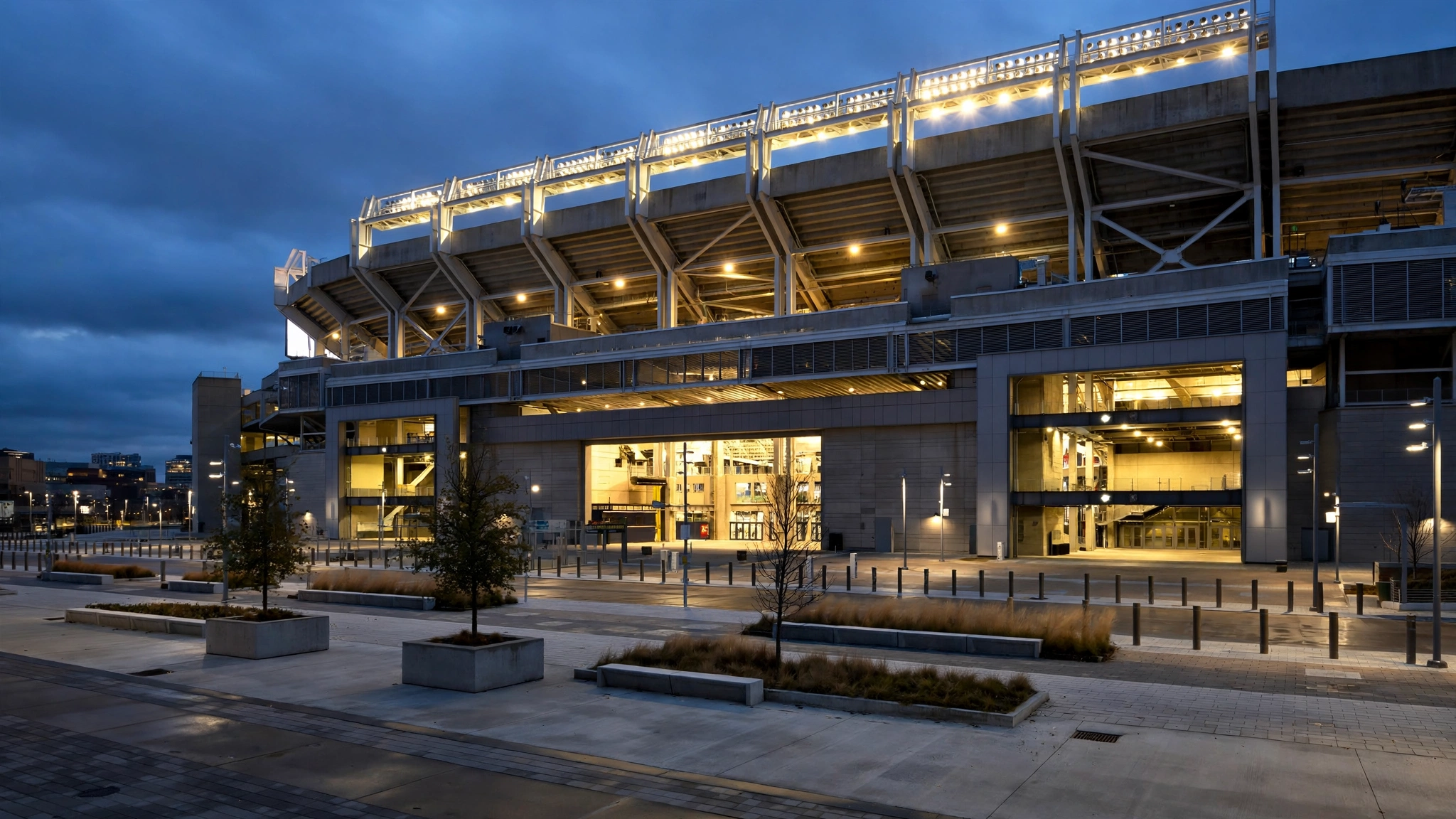 A football stadium exterior with lights on at dusk and an empty plaza in front