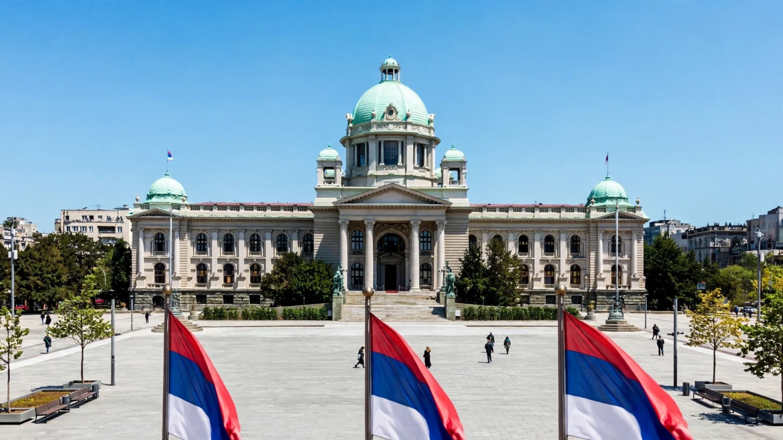 The parliament building in Belgrade seen across a public square, with red, blue and white flags in the foreground and people walking in the square.