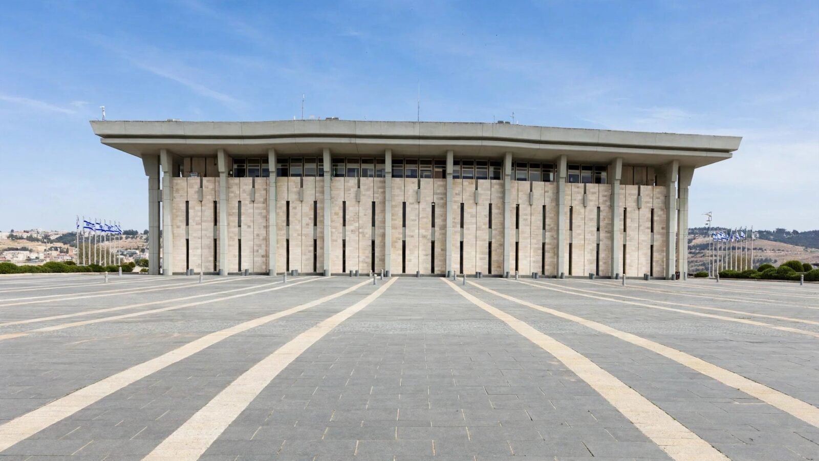 The Knesset building in Jerusalem, a wide stone parliament building with flag poles on either side, under a clear sky