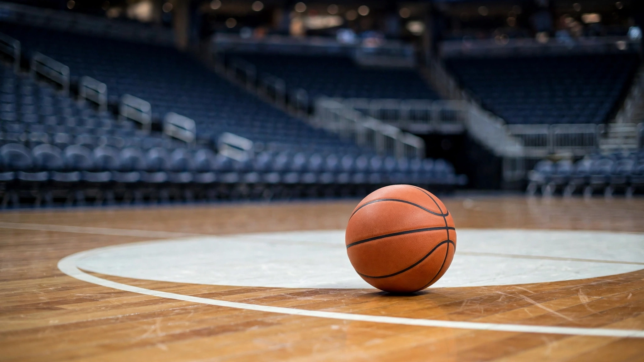 A basketball resting on a polished wooden court near the center circle with empty arena seats behind it