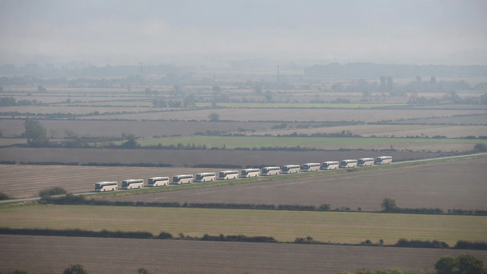 A line of white coaches travels along a road through flat farmland