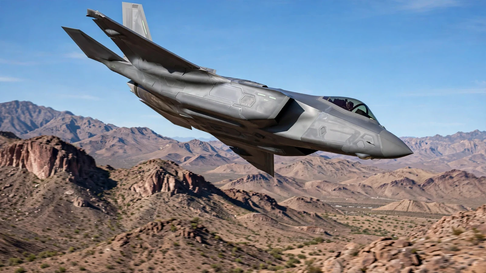 A grey stealth fighter jet banking in flight over desert mountains under a clear sky.