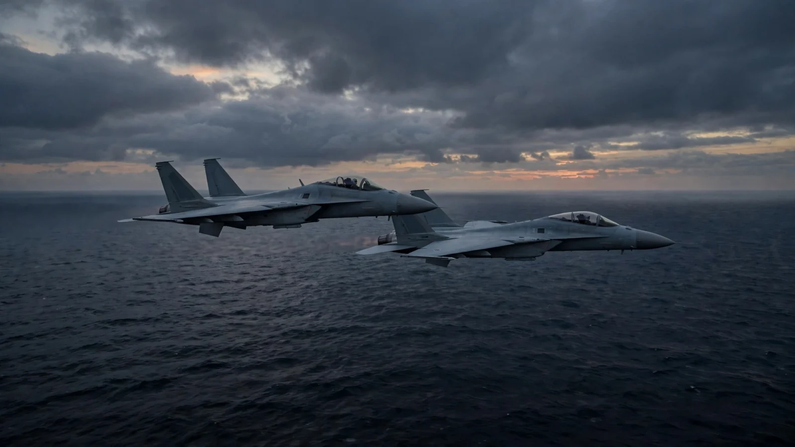 Two grey fighter jets flying in formation over a dark sea at dusk under a cloudy sky.