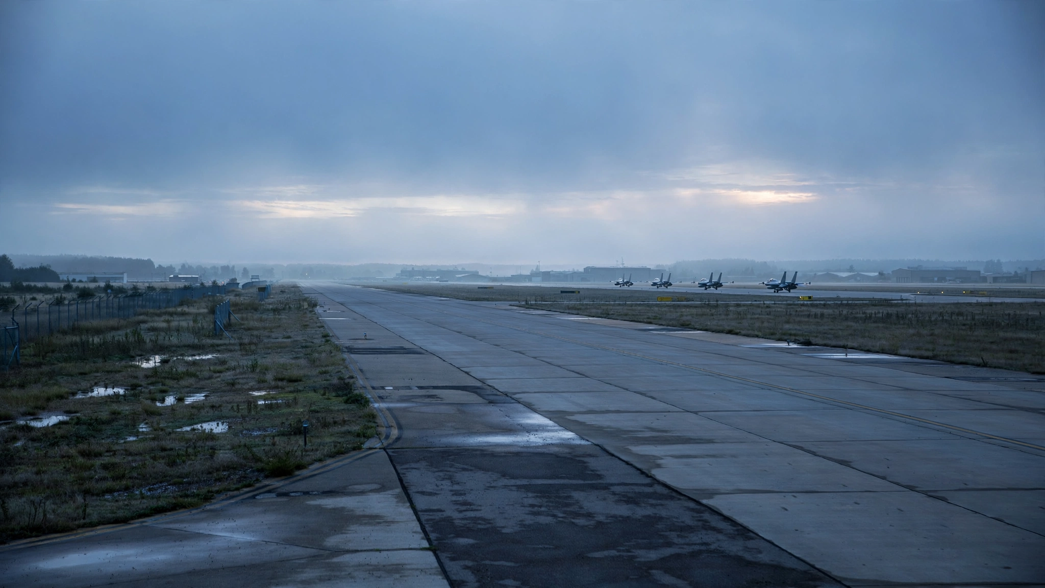 A military air base runway at dawn with fighter jets parked in the far distance.