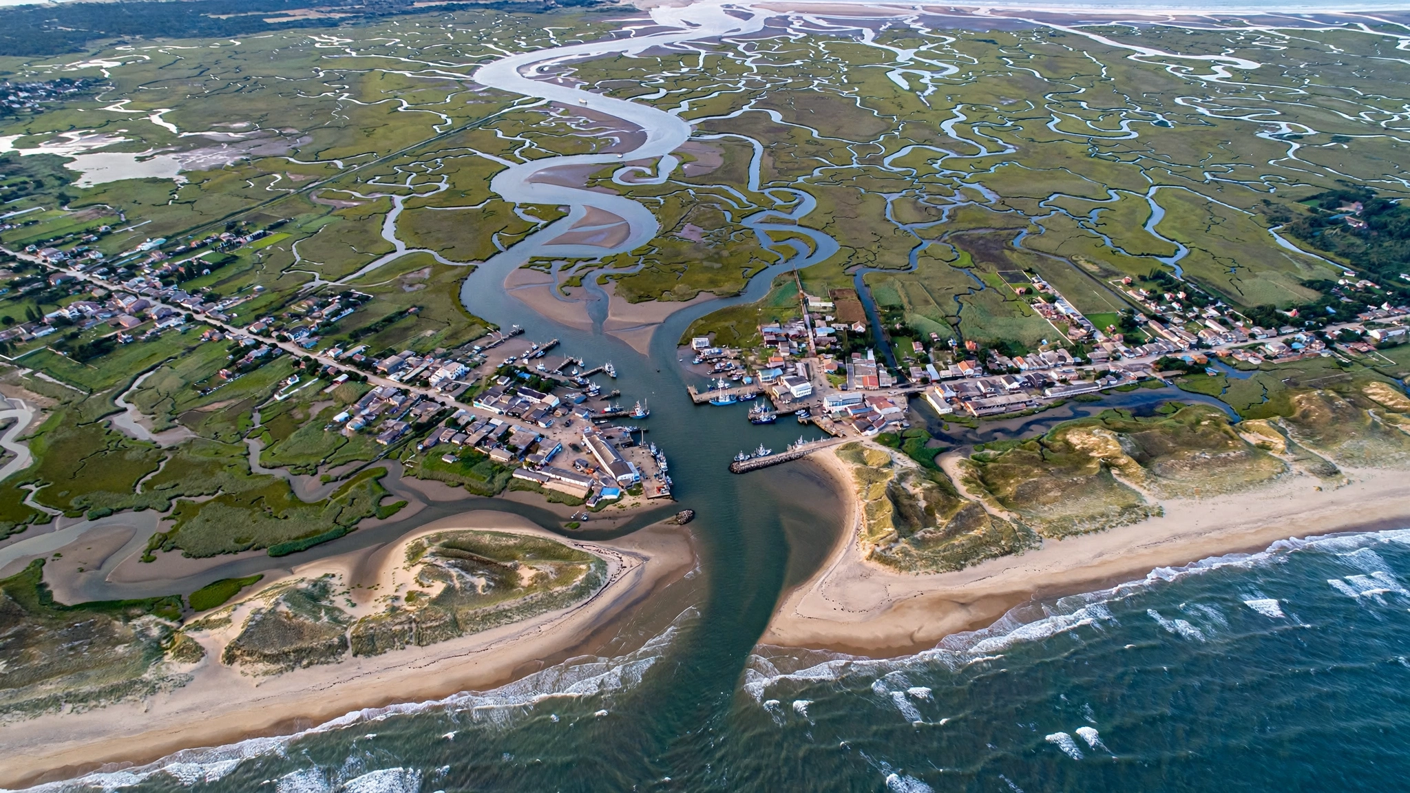 An aerial view of a coastal town at the mouth of a river delta meeting the sea, with green marshland and sandy beaches.