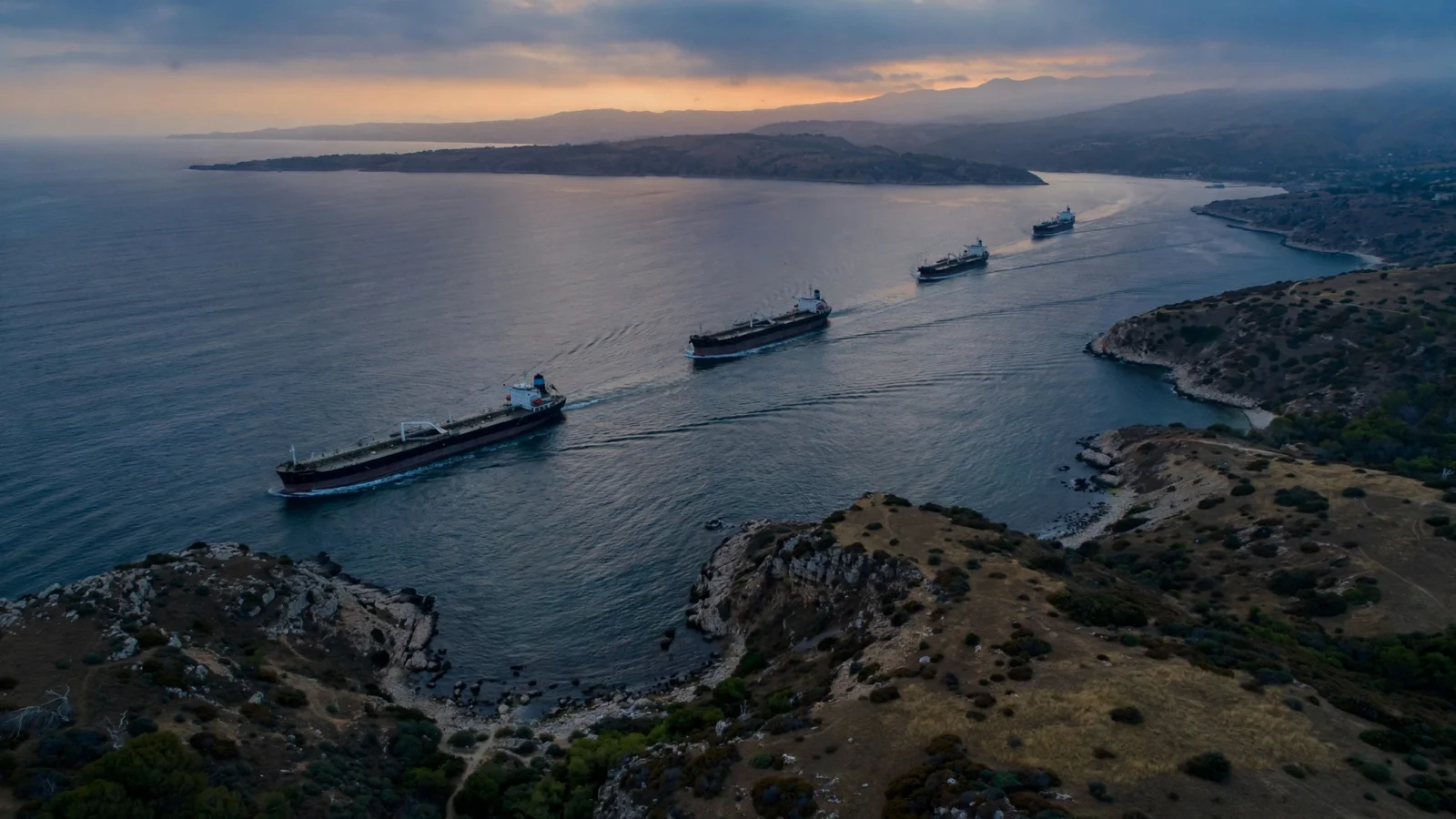 Large oil tankers crossing a narrow sea strait at dusk, with a coastline on the horizon.