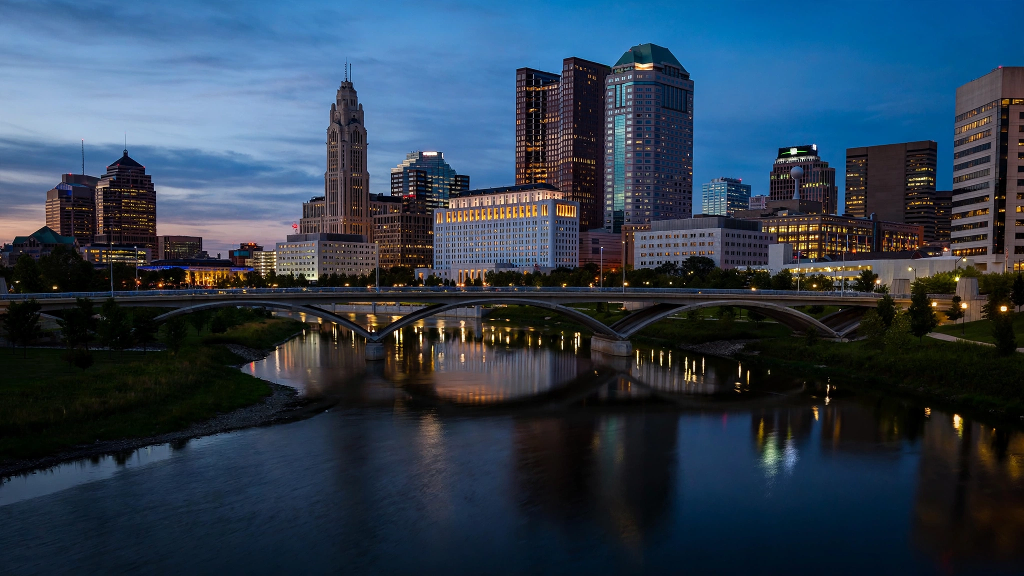 The downtown Columbus, Ohio skyline at dusk seen across a river with a bridge