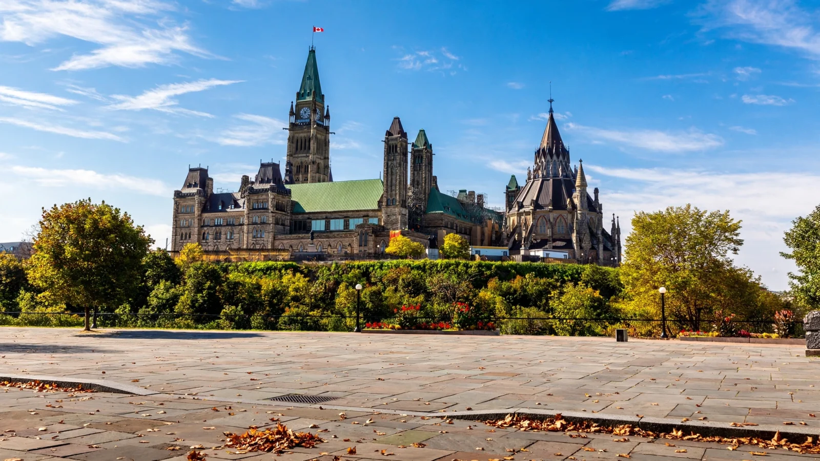 Parliament buildings in Ottawa with a flag on the tower, seen across a paved plaza with trees in autumn color