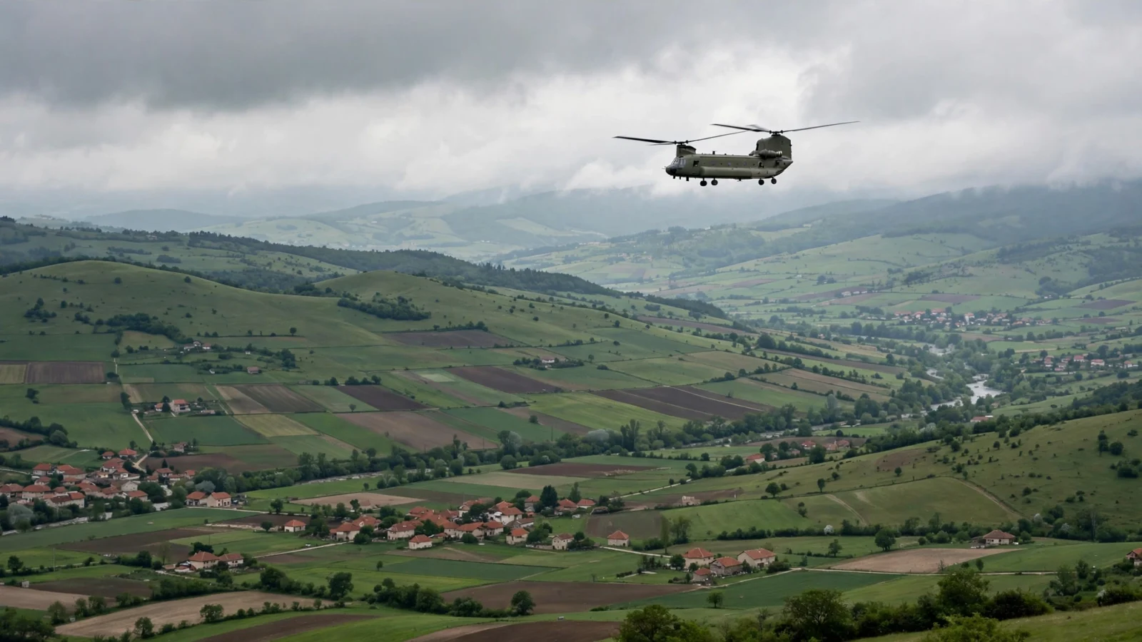 A twin-rotor military transport helicopter flying over green hills and farmland under a cloudy sky.