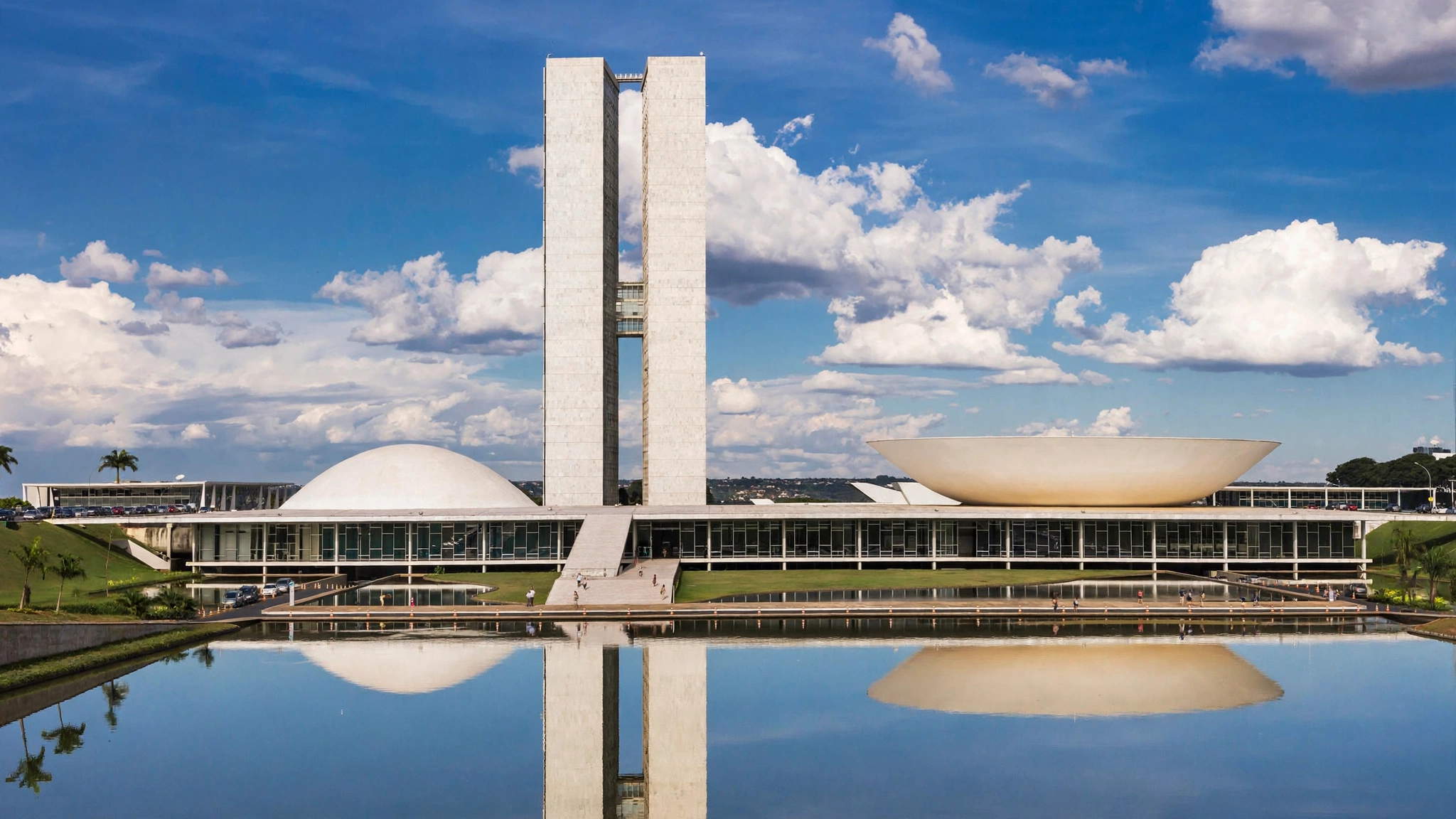 The white National Congress building in Brasilia with two towers and curved roofs, reflected in a pool of water under a blue sky