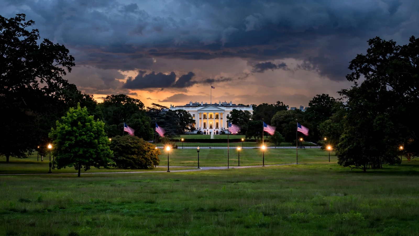 The White House exterior at dusk with lit windows, American flags on poles across the lawn, and glowing street lamps under a cloudy evening sky.