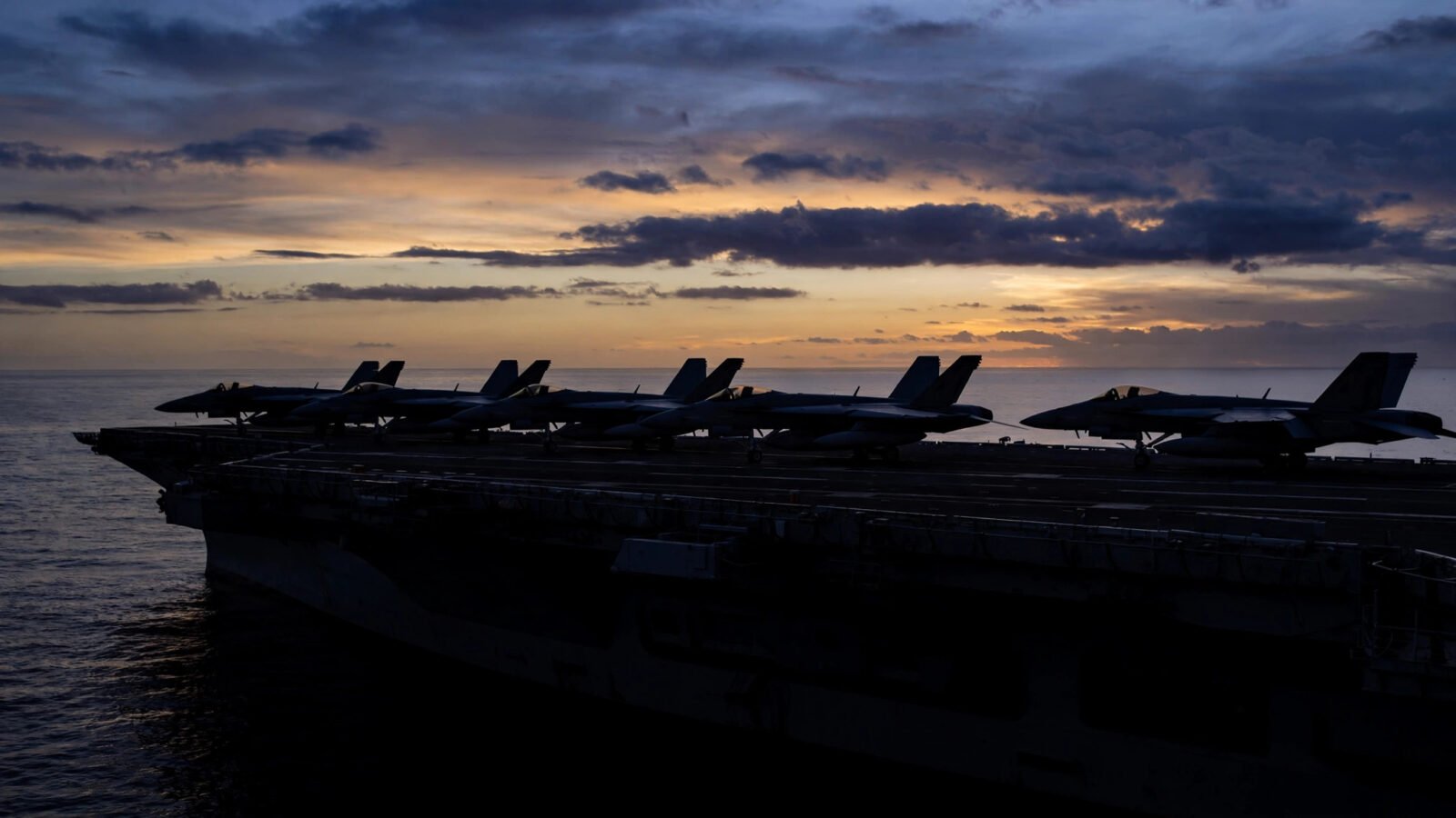 Silhouettes of fighter jets on an aircraft carrier flight deck at dusk over the ocean.