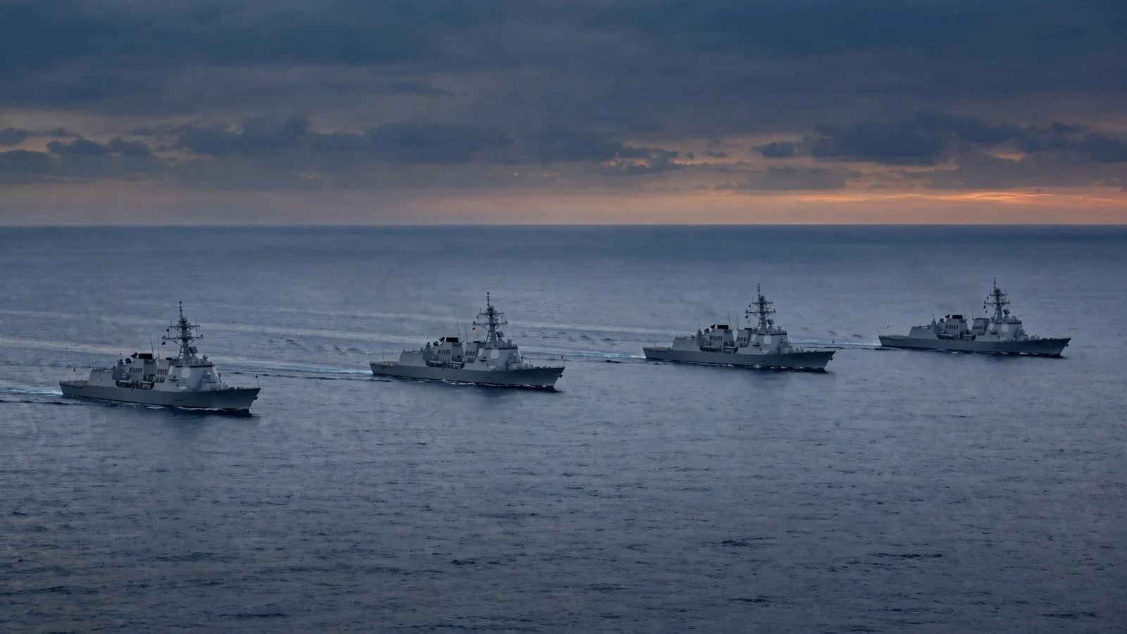 Four grey naval warships sailing in formation on open ocean at dusk