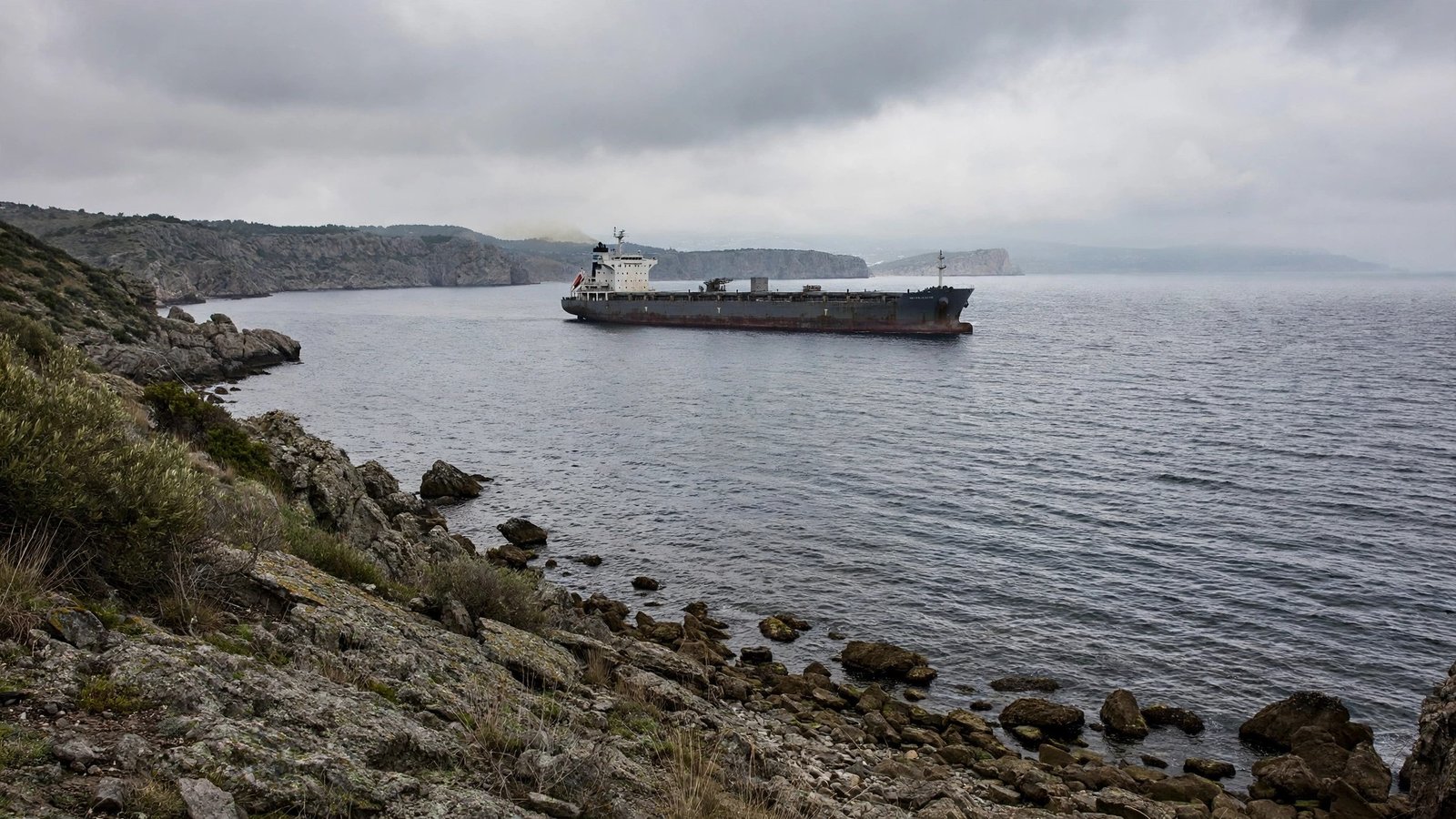 A cargo ship sailing past a rocky coastline under a cloudy sky