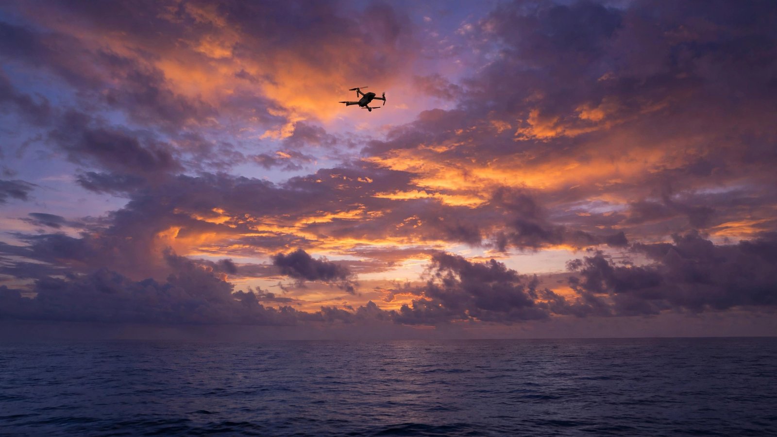 A small drone silhouetted against orange and purple clouds over the sea at dusk