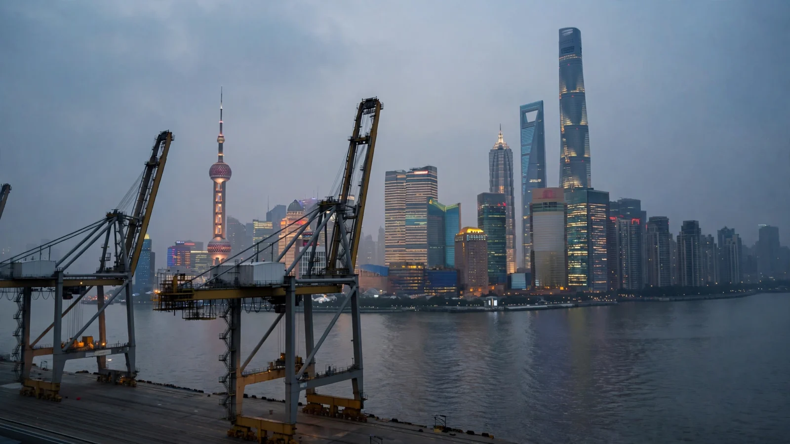 The Shanghai skyline at dusk seen across the water, with container cranes in the foreground.