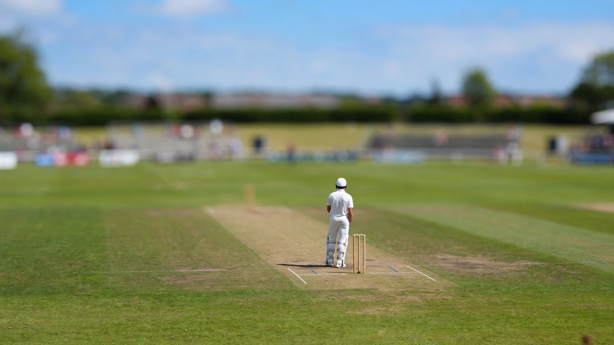 A cricket player in white kit standing at the crease, seen from behind, on a grass field.