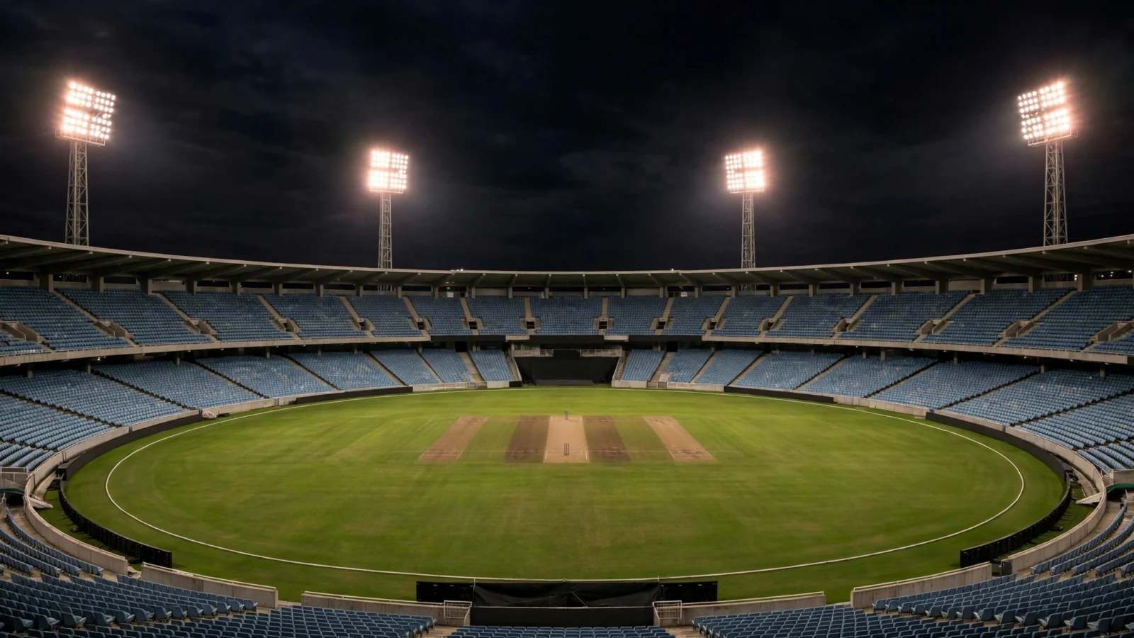 An empty cricket stadium under floodlights at night with a green outfield and a pitch in the center.