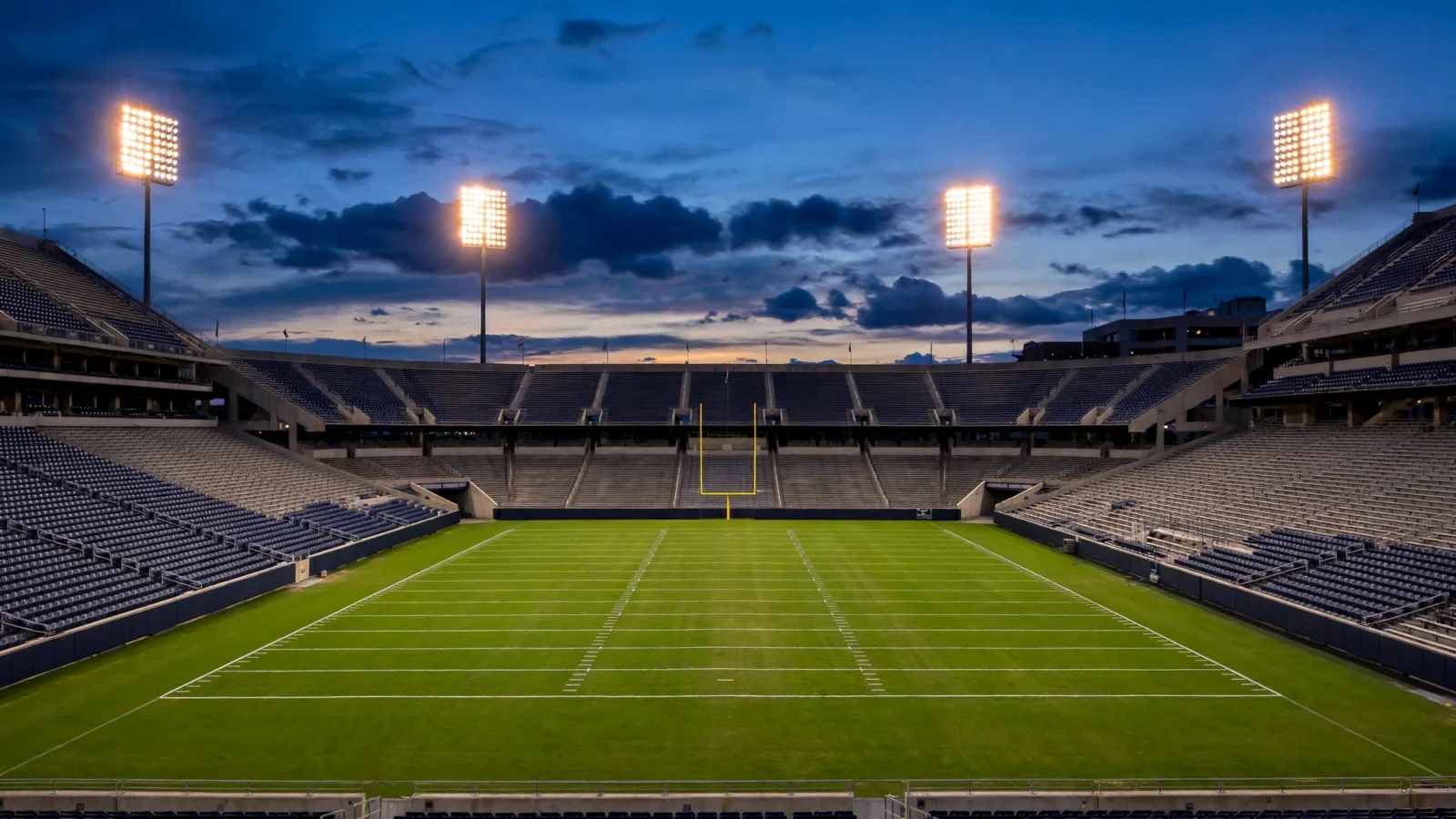 An empty college football stadium at dusk with floodlights on over a green field and goal posts.