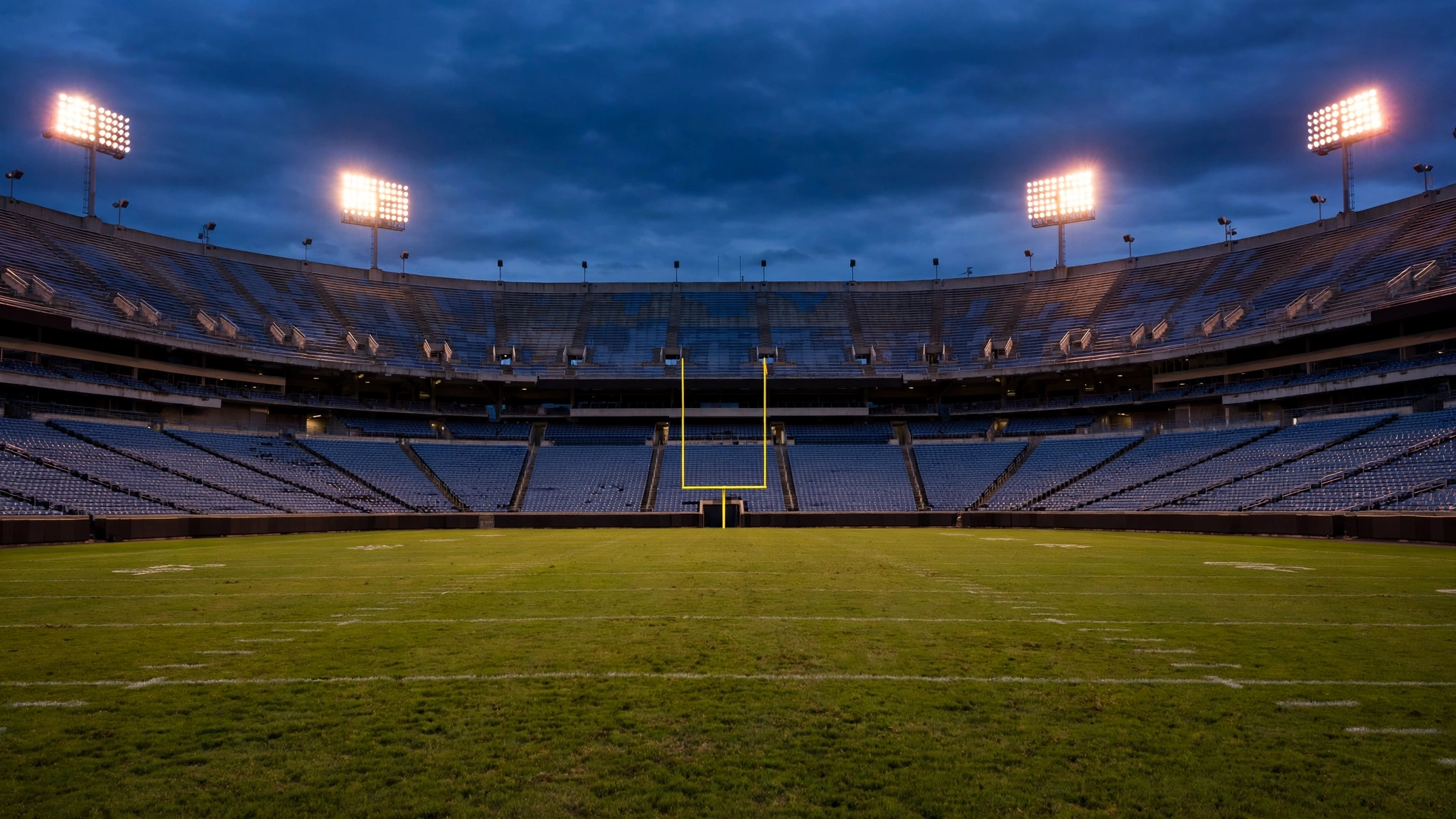 Yellow football goalposts on a grass field in a large stadium under floodlights at dusk