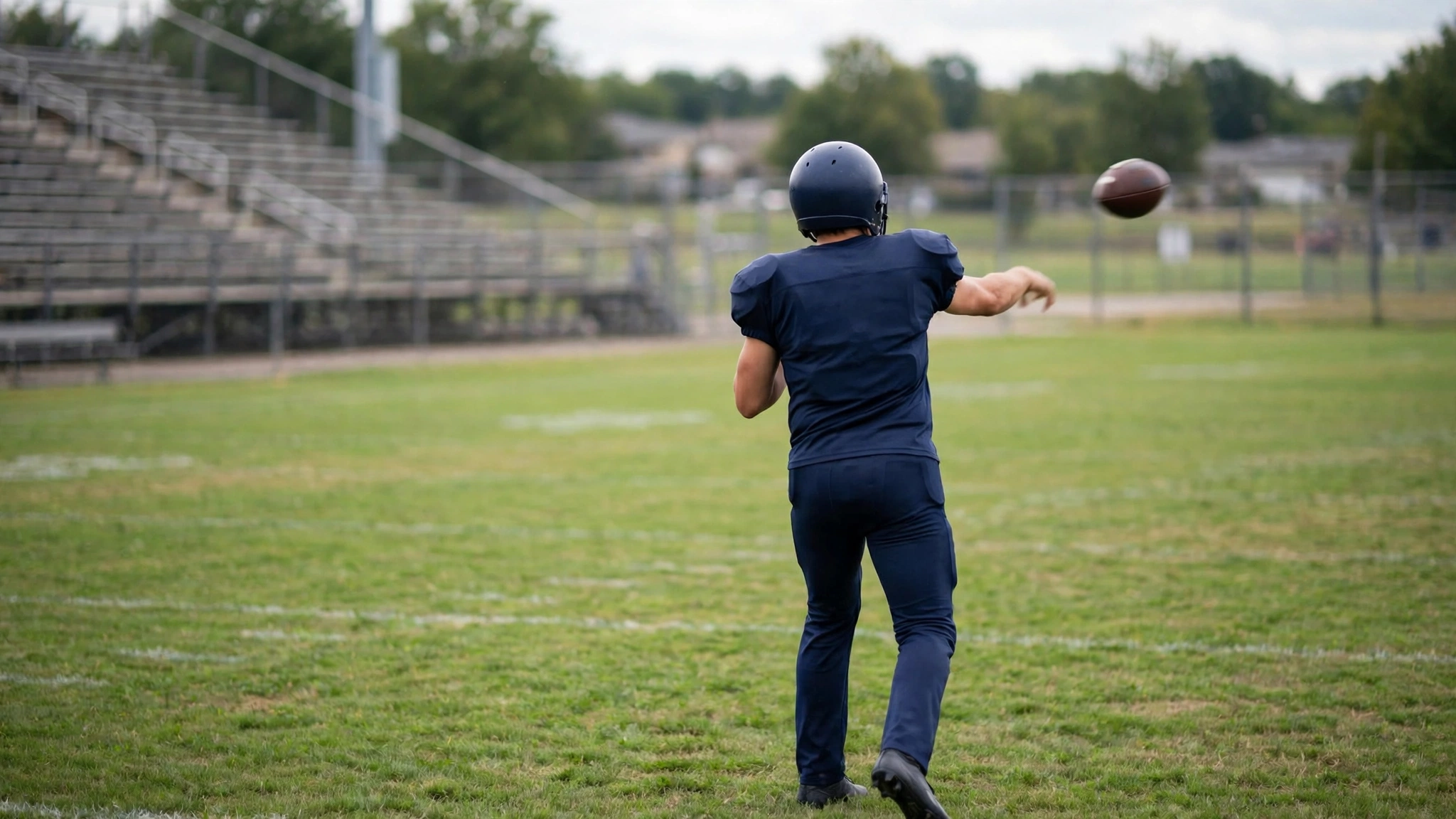 A football player in a plain dark uniform throwing a pass on a grass field, seen from behind