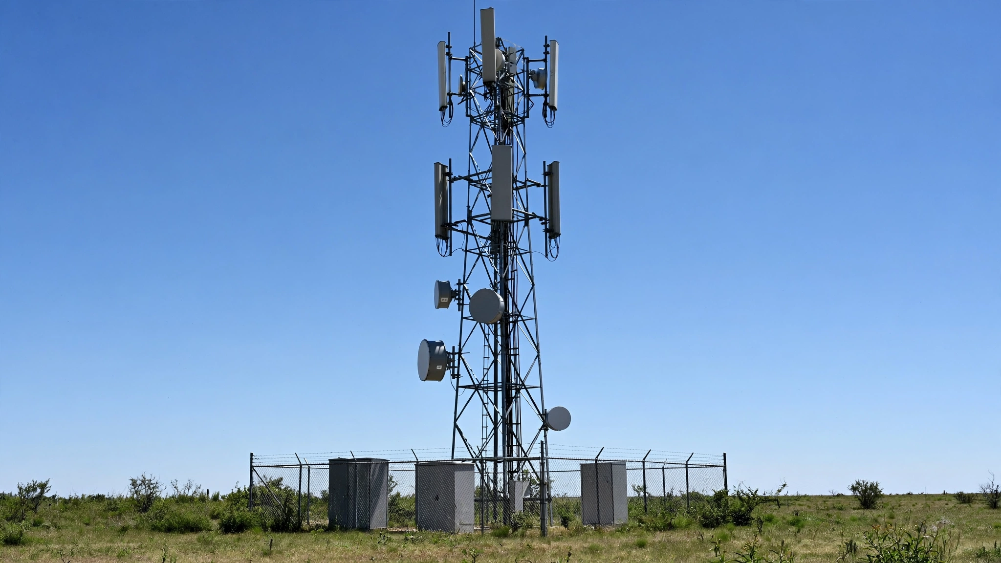 A metal cell phone tower with antennas standing in an open field under a clear sky