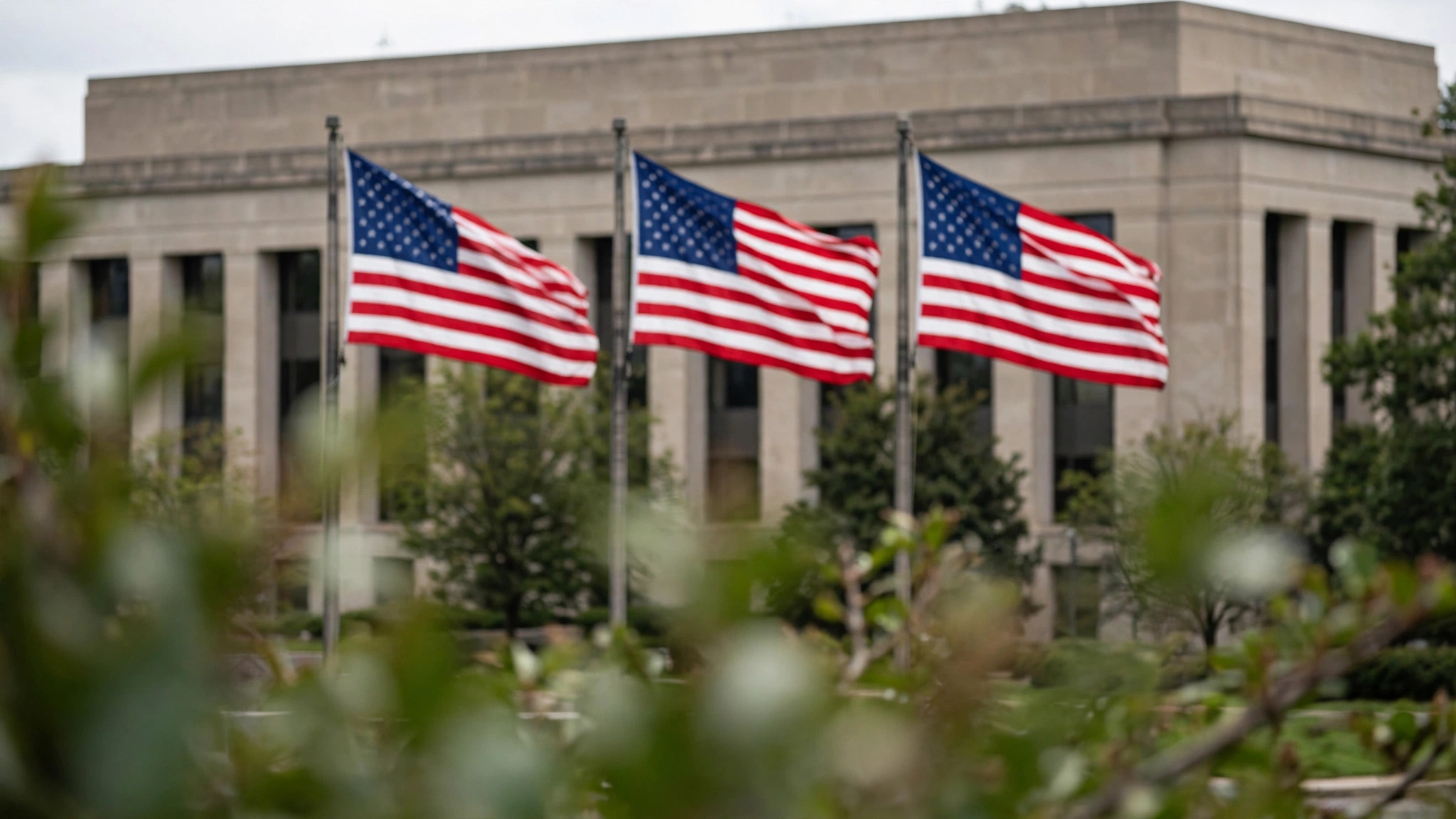 Three American flags flying on flagpoles in front of a plain stone government building.