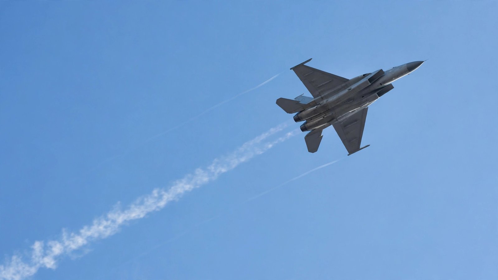 A fighter jet flying high in a clear blue sky with faint contrails
