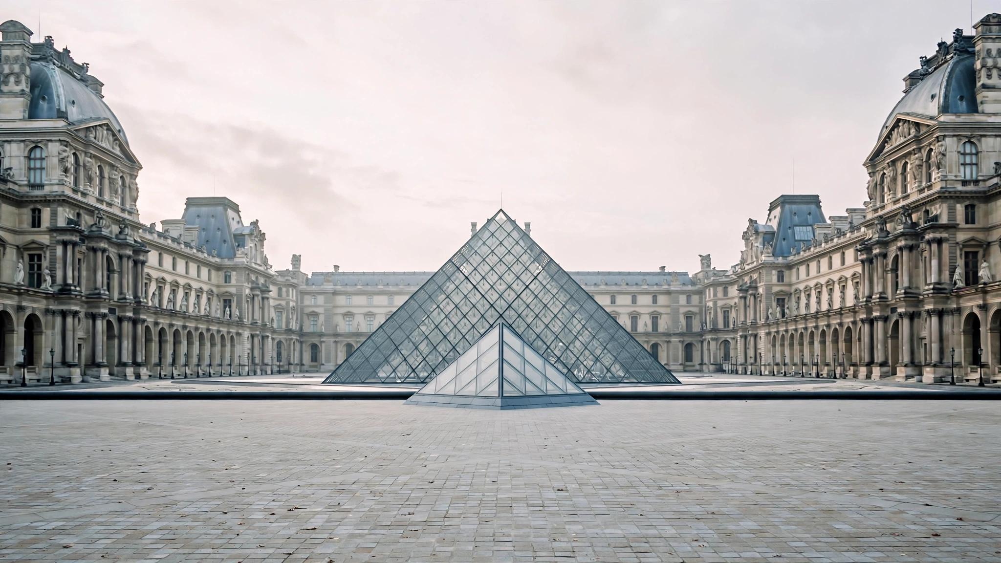 The glass pyramid of the Louvre museum in an empty courtyard under a pale morning sky.