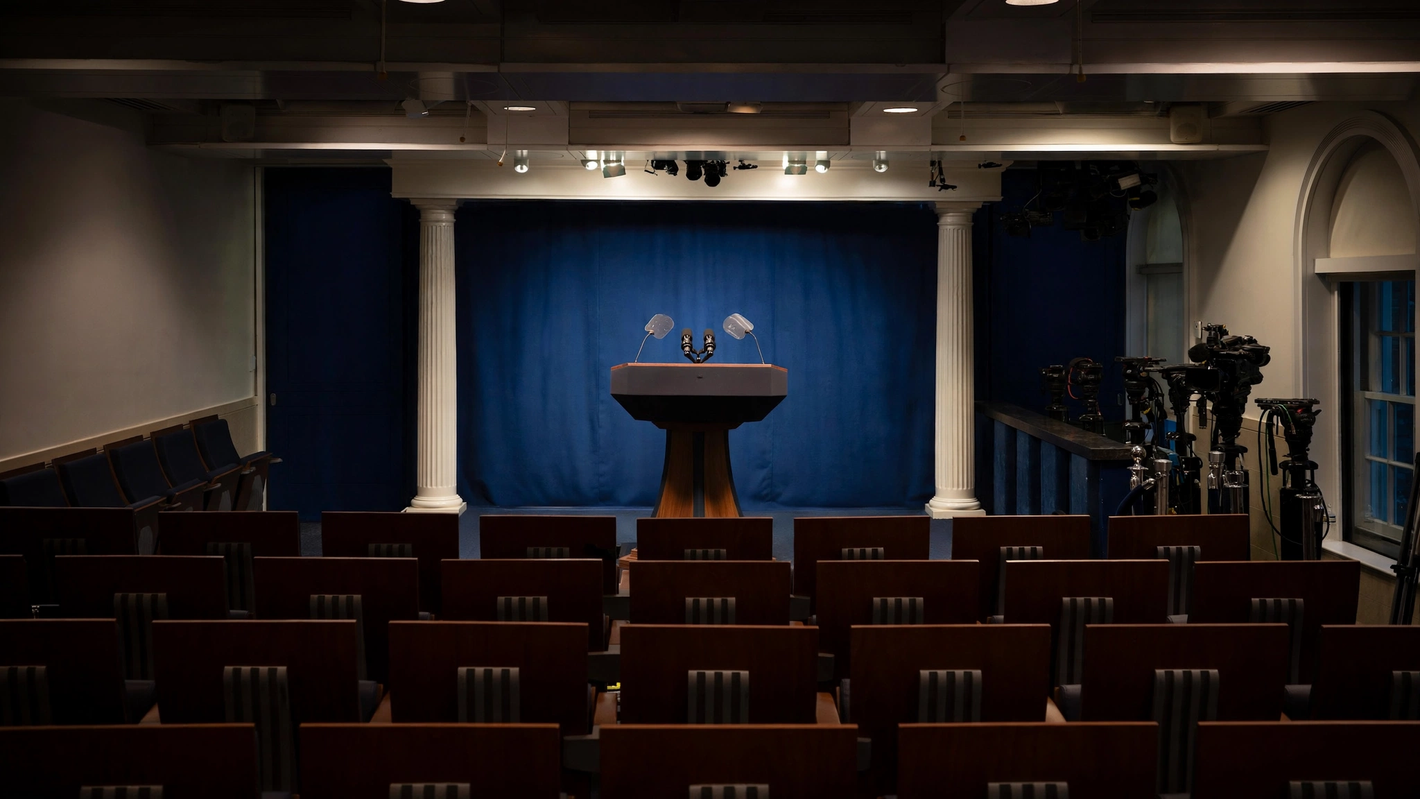 An empty White House press briefing room with a podium, microphones and rows of chairs in front of television cameras.
