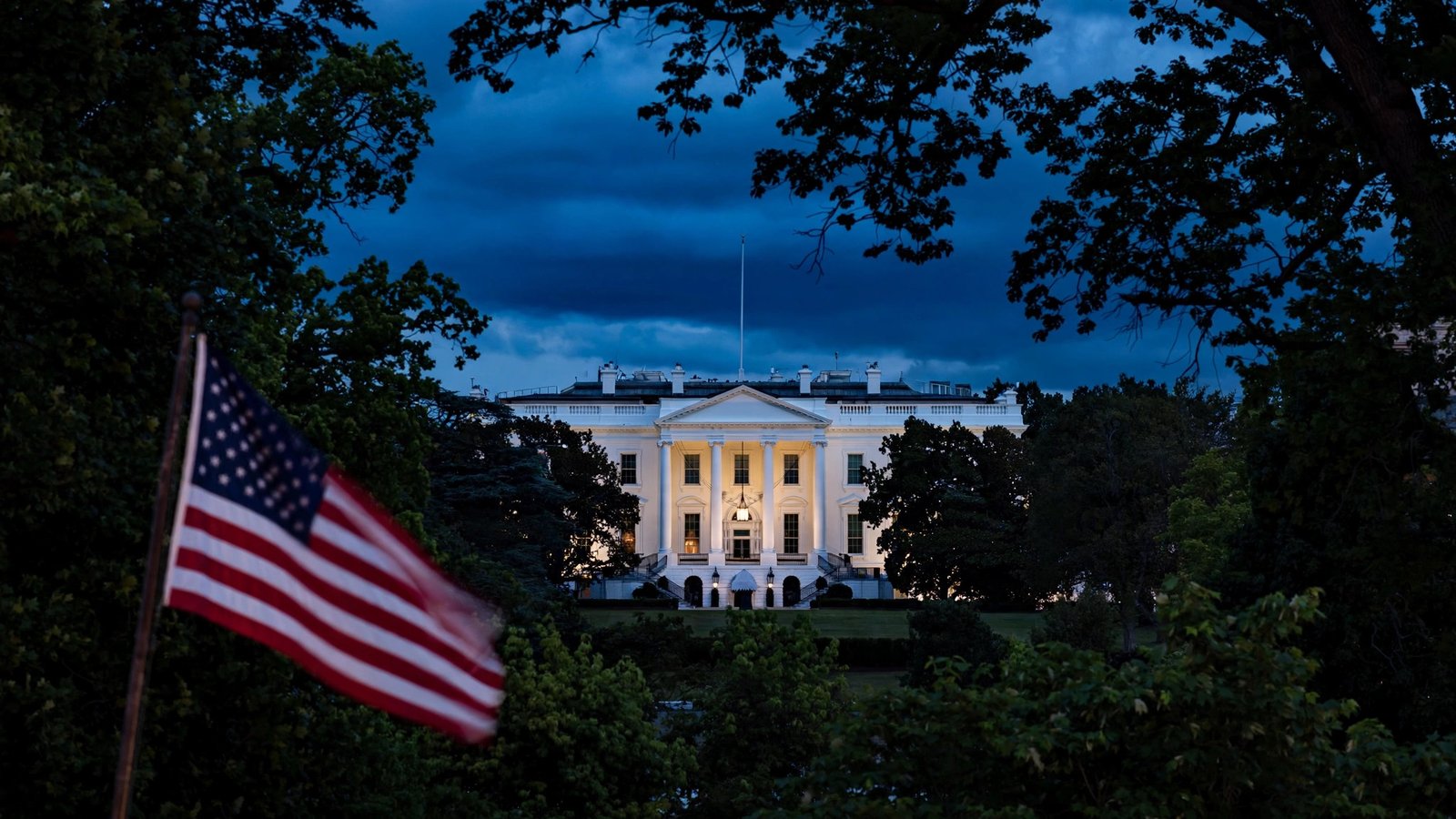 The White House at dusk with an American flag in the foreground, framed by trees.