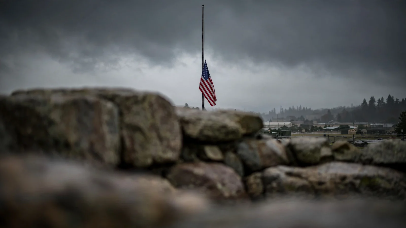 An American flag flying at half-mast under dark storm clouds above a stone memorial wall at a U.S. Army installation.