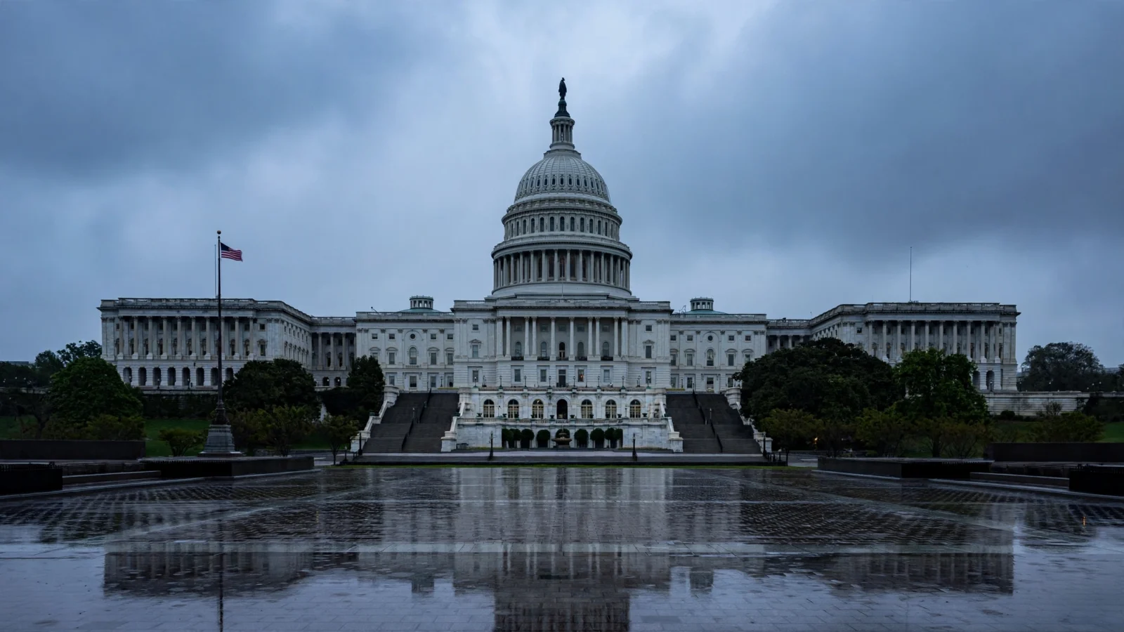 The United States Capitol building in Washington, D.C., seen from across a wet plaza under a gray overcast sky, with an American flag flying to the left.